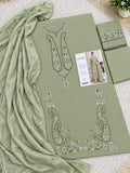 An olive green traditional Pakistani shalwar kameez suit set with matching dupatta, featuring intricate silver and white embroidery on the chest and hem, laid out on a white textured rug with a brand card and a small plant pot.