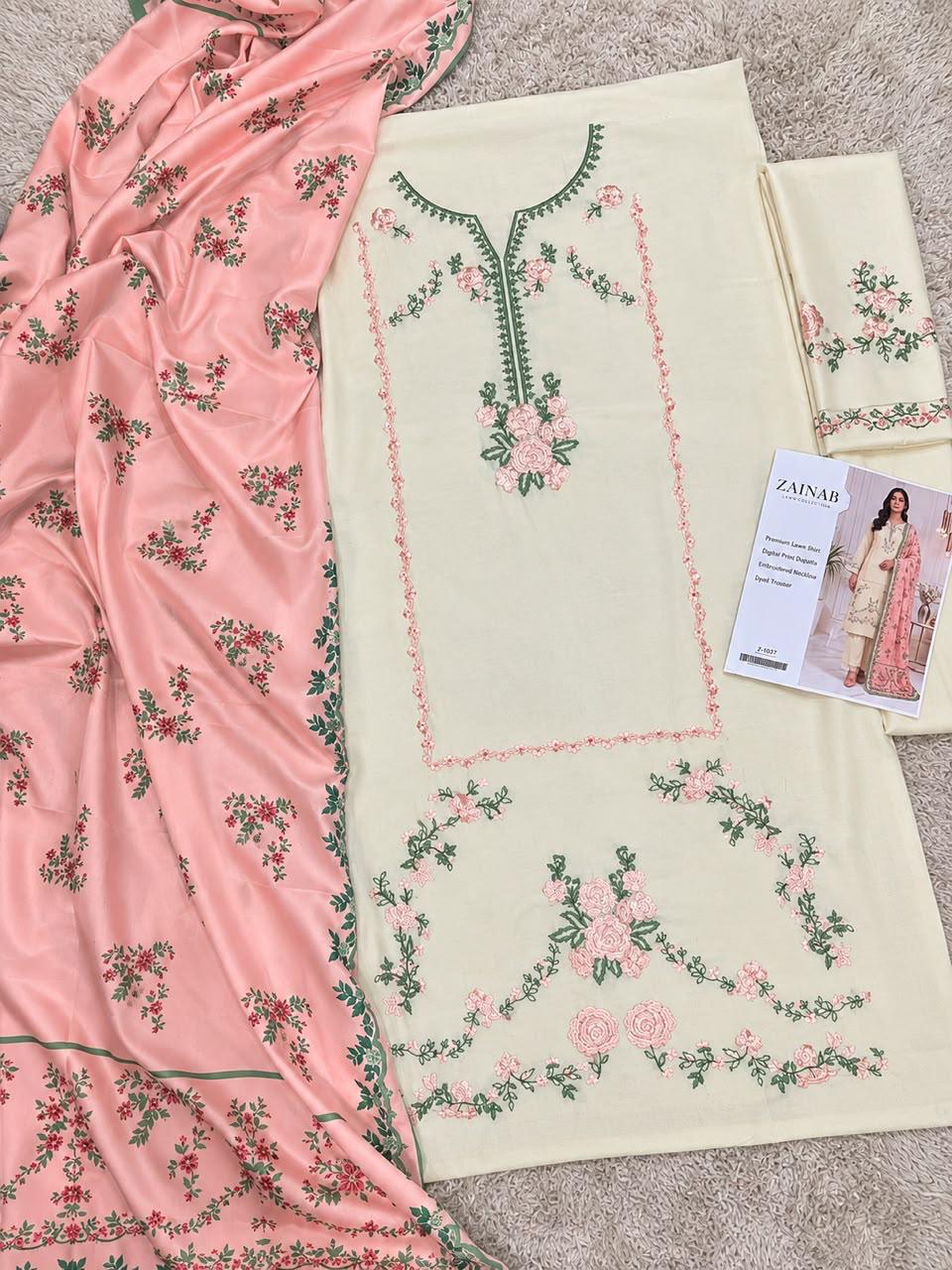 A flat-lay view of a traditional South Asian salwar suit set, featuring a cream-colored kurta with green and pink floral embroidery, a matching pink dupatta with green and pink floral patterns, and a folded matching fabric piece. A brand card for 'Zainab' is visible on the right side.