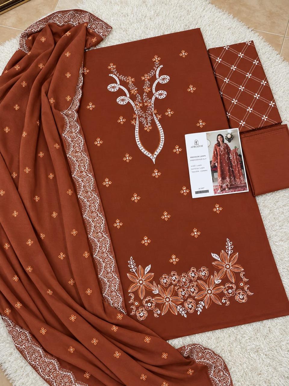 A rust-brown salwar kameez suit set laid flat on a white textured rug, featuring a printed fabric piece with white and gold floral embroidery, a matching printed dupatta with white lace borders, a folded patterned fabric piece, and a small printed card.