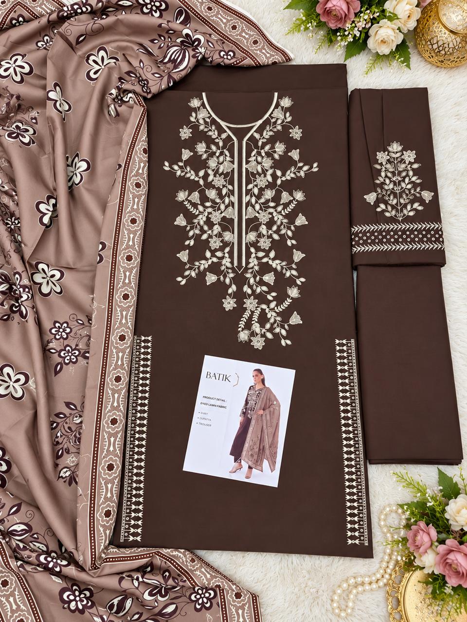 A flat-lay presentation of a brown three-piece salwar kameez suit set, featuring a kurta with intricate white floral embroidery, matching trousers, and a dupatta with matching floral patterns. A small brand card is visible on the kurta.